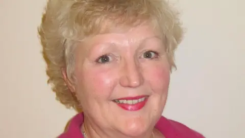 Jane Slatter Jane Slatter with medium-length light brown hair, wearing red lipstick and a pink top and smiling at the camera. There is a light-coloured wall behind her.