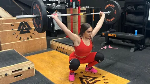 Arzu Dutta/BBC Annie in a squat position, holding a weighted barbell over her head. She is wearing a red gym set and training for the IWF Youth World Championships.