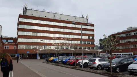 Anttoni James Numminen A five-storey red brick building with windows along the width of each floor and the words York Hospital Main Entrance written on it in white lettering. I the foreground is a car park full of cars.