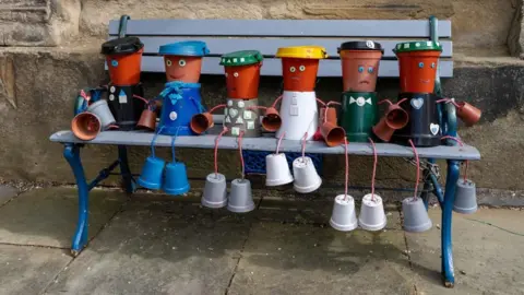 Alan Thornton Six figures made from flower pots sit on a bench. Each has two small flower pots for feet with strings for legs attached to a large coloured pot for the body with a smaller brown pot decorated with eyes and a mouth on each.