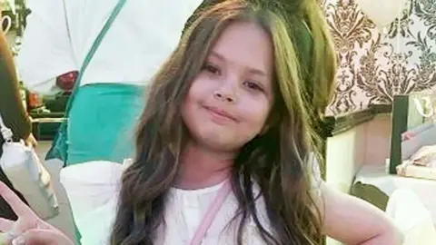 Olivia has long, wavy brown hair and is wearing a white t-shirt; she is smiling at the camera.