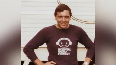 East Suffolk and North Essex NHS Foundation Trust An old photo of John Alborough as he smiles at the camera. He wears a brown long-sleeved top with the words Hospital Radio Ipswich on it as well as an emoji-style face with radio headphones on. John has brown hair and rests his hands on his hips.