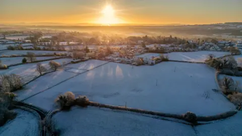 Anna Barclay An aerial image showing Holcombe village and the surrounding fields at sunrise on a frosty day. There is a thin layer of snow on the ground and the image looks cold and misty.