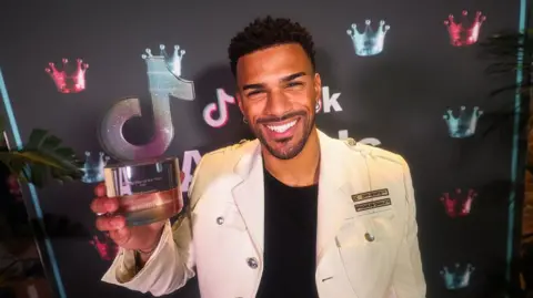 Getty Images A man in a white embellished jacket smiles at the camera, holding a sparkly glass award in the shape of the TikTok logo.