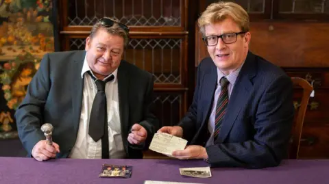 Bearnes Hampton & Littlewood The image shows Dean Buffini wearing in a dark suit with black tie, which is loosened and dark glasses on his head. He is sitting besides Brian Goodison-Blanks wearing a navy suit and glasses who is holding a gun licence issue to Ronnie Kray in 1964.
There is a purple cloth on the table in front of them. Mr Buffini is holding a ring and there is a colour photo featuring his step-father Charlie Kray in front of him and a black and white photo to his right.