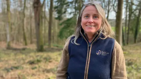 Fiona Irving / BBC A woman with blonde hair down to her shoulders looks at the camera. She has a pair of glasses perched on her head and is wearing a blue and brown zipped fleece. The jumper has a brown logo with the words 'English Woodlands Forestry' next to it. In the background are trees.
