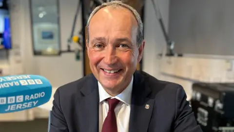 A mid shot of Deputy Lyndon Farnham. He has very little hair and is smiling into the camera. He is wearing a suit, with a maroon tie.
