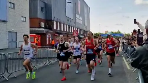 A large group of runners taking part in a road race, moving past metal barriers outside a stadium. Spectators line the right-hand side with some holding up phones to record the runners as they pass.