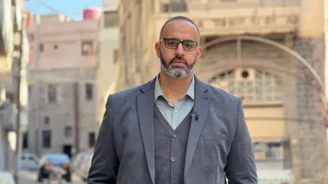 Francesco Tosto/BBC Man in suit and glasses stands outside in street looking at the camera