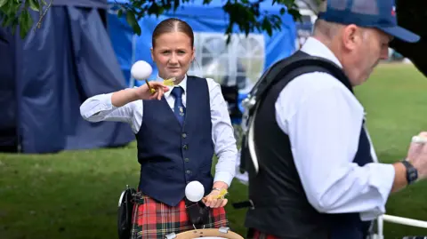Pacemaker Press A young woman is playing drums. She's wearing a red checked kilt and a navy waistcoat. A ,am wearing a cap is next to her. 