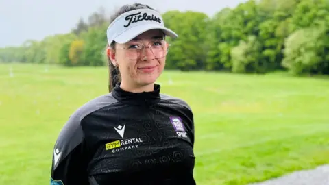 Amateur golfer Robyn Fowlie smiling at the camera at a golf driving range, she is wearing a branded golf hat.