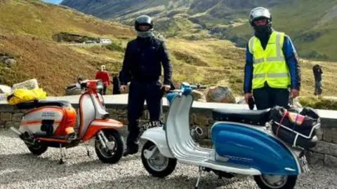 Lambretta Club Scotland Two men standing with their Lambretta scooters with hills and mountains behind them. They are both wearing helmets and is wearing a high-vis jacket.