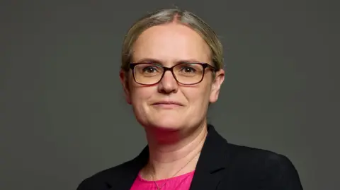 UK Parliament Lizzi Collinge looking directly into the camera. She has blonde hair, parted just off-centre, and is wearing glasses and a black blazer with a pink top.