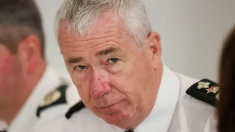 Chief Constable Jon Boutcher wearing a white shirt with psni shoulder titles. Behind him is another police officer in a white shirt. 