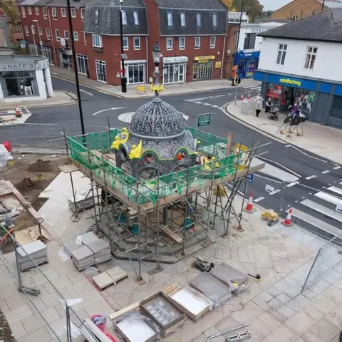 Mallets of March A view of the Coronation Fountain in March town centre from a third storey window. The metal structure is surrounded by scaffolding, and a mini roundabout is behind it.