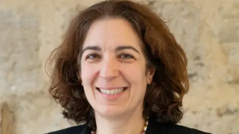 University of Oxford Head and shoulders shot of a smiling Prof Mina Fazel who has shoulder length brown hair and is wearing a black jacket.