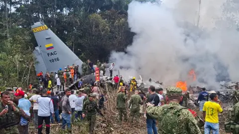A crowd of people, including army personnel, gather at the crash site. Some stand on the wing on the wrecked plane. Smoke rises and fires burn out of the wreckage.