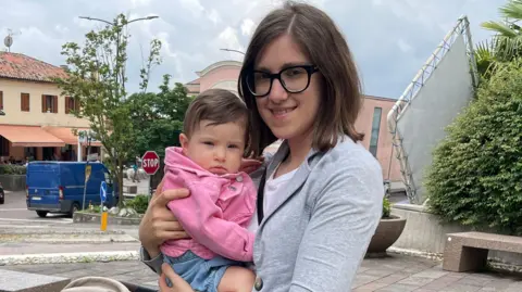 Valentina, a woman with dark hair and glasses, holds a baby in a pink jumper