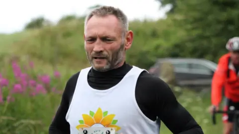 A close up image of Gary McKee running. He has a grey beard and is wearing a black long sleeved top with a running top with a sunflower logo 