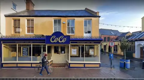 Google A person pushing a bicycle and wearing blue jeans and trainers, a dark jacket and a red woolly hat, walks past a cafe with a large sign reading Cafe CoCo. The walls of the building are painted yellow.