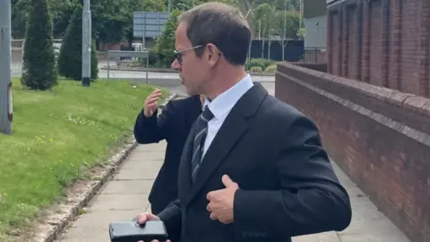 Dr Isles is wearing a suit  and tie as he leaves the court. He is captured in a side-on view and is looking away from the camera. He has short brown hair and glasses on.