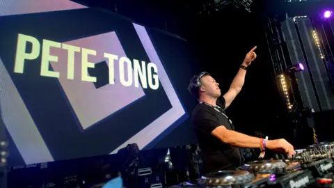 Pete Tong' mixing music with one hand in the air and a hige screen with his name on it behind him