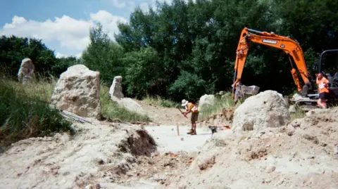 Workmen wearing high vis jackets with a digger working on what looks like a quarry but is in fact the start of a new henge. Large chalk stones form a circle and in the middle a workman is hammering something. There are trees in the background.