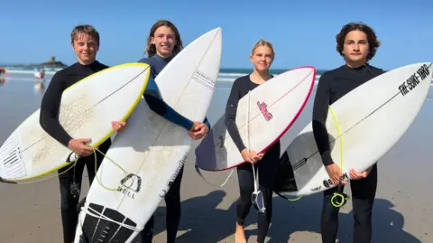 Four young surfers in black wetsuits stand on a beach holding surfboards. The ocean and blue sky form the backdrop, with the group facing the camera on wet sand near the shoreline.