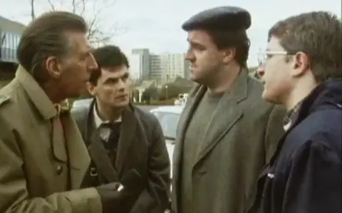 Ricky Fulton, Gerard Kelly, Iain McColl and Joe Mullaney stand in a row talking. They are all wearing heavy overcoats and McColl, the tallest and biggest of the men, is wearing a flat cap. A large concrete building can be seen in the background.