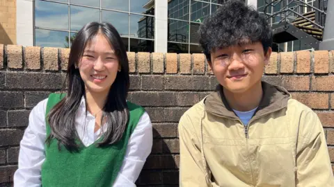 Sunwoo and Sangweon smile at the camera. Sunwoo has long black hair and is wearing a white shirt. Sangweon is wearing a beige vip up jacket and has short black hair. They are seated with a brick wall behind them.