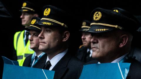 Aer Lingus pilots who were protesting in Dublin over their pay. There are two male pilots in the foreground, wearing their caps, holding placards, with three pilot in the background