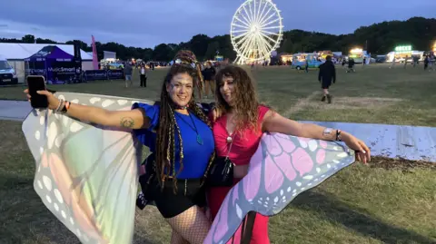 Two women stand in a field at dusk. They are sharing a large fancy-dress set of butterfly wings that they are stretching out around them. The woman on the right has dreadlocks and a blue top. The woman on the left has wavy brown hair and a pink top. There is a ferris wheel and stalls in the background.