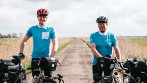 Joshua and George Kohler, both wearing cycle helmets, pale blue cycling tops and black cycling trousers. They are standing by and holding their bikes, which are laden with black panniers, and are pictured on a track in Norfolk, with flat fields either side