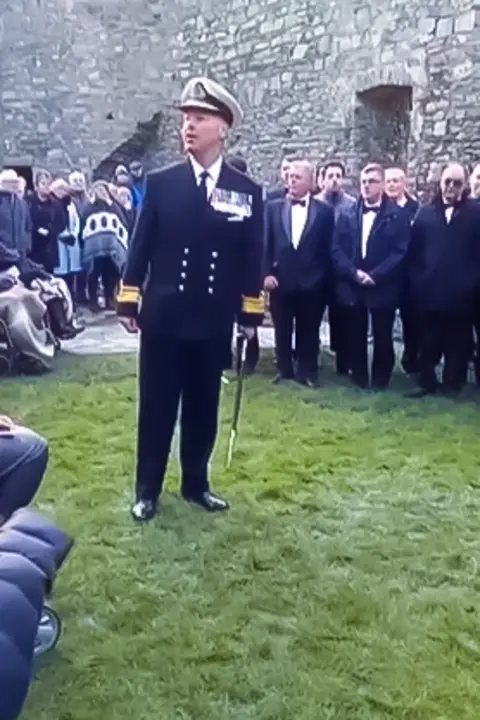 A man in a navy officer's white hat and blue blazer stands addressing a crowd against a castle wall. In his left hand he holds a ceremonial sword.