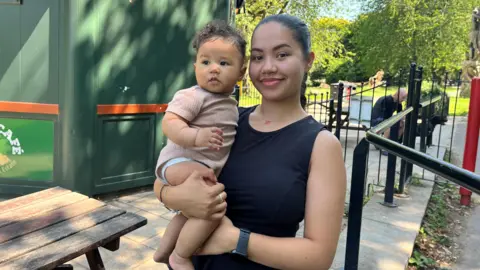 Aaliyah standing in a Bristol park as she holds her baby in her arms. She is wearing a black dress and has dark brown hair tied back in a ponytail. She is smiling at the camera,