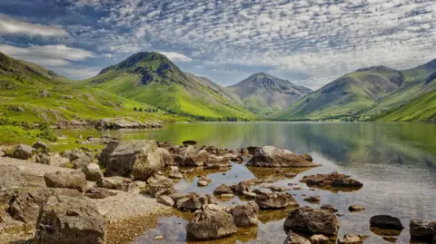 Getty Images Reflections of foreshore rocks in the clear water of Wastwater. Green mountains stretch out on the far side of the lake.