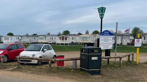 View of the sparsely populated North Beach car park at Heacham. There are three cars parked on the sand next to a parking metre and a yellow barrier limiting the height of cars that can be parked there. 