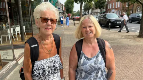 Two women wearing back packs stand in a street looking at the camera.