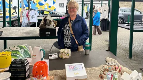 Jessica Bayley/BBC A woman stands behind a market stall, she has short blonde hair and glasses and is wearing a navy waterproof coat.
