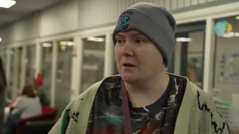 A person wearing a black patterned t-shirt, green cardigan and a grey woolly hat is stood inside the community centre.