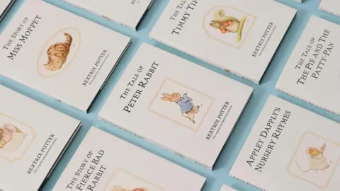 Penguin Random House UK Lots of Beatrix Potter books arranged on a table. The books all have white covers with a character drawn on them and the title of the book, such as The Tale of Peter Rabbit and The Story of Miss Moppet. The table they are on is baby blue. 
