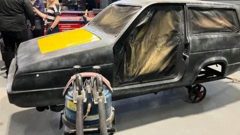 Graham McKechnie Grey three-wheeled car with yellow rectangle on the bonnet. The seats have been covered with gold-coloured sheeting. The wheels have been removed. There is a blue circular machine beside the car. People are visible behind the car.