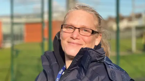 A woman with blonde hair stands smiling for a photograph. It is a bright day and the sun is shining on her face, lighting it up. She is wearing rectangular black glasses, a thick black coat and has a lanyard with 'staff' written on it around her neck. The background is blurred but she appears to be standing in a sports area/training ground as there is fencing and grass behind her.