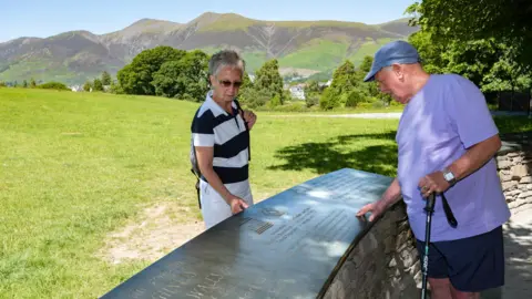 A man and a woman look at the UNESCO world heritage plaque at Crow Park in Keswick. The plaque is a a piece of slate with an inscription, mounted on a drystone wall, to look like a long table. Behind the two people, are the fells in the background.