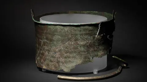 David Brunetti/National Trust A photo of the Bromeswell Bucket which has been displayed on a black surface with a black background. Part of the bucket is missing and etchings can be seen on the bucket's surface.