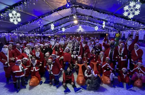 A large group of people of all ages dressed as Santa pose for the camera on the ice rink at Clarks Village in Street in Somerset