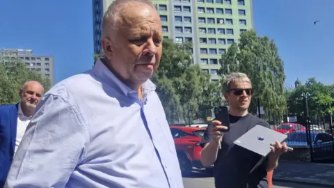 John Stiles talks outside the court.He wears a blue open-necked shirt and is balding with grey. There are journalists recording his statement. 