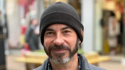 Man with dark and grey beard wearing a black beanie hat.