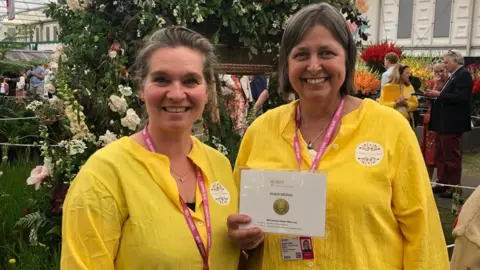 Two women stand next to each other, smiling at the camera. They are both wearing bright yellow blouses and pink lanyards. The woman on the left has brown and grey hair pulled back into a ponytail. The woman on the right has bobbed brown and grey hair and is holding an award with the words "RHS" and "GOLD MEDAL" on it.