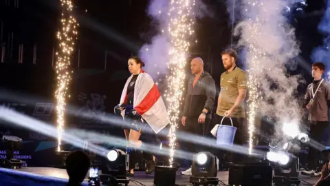 Andrew Cleminson Alanna Pritchard walks on a stage with lights and fireworks. He is wearing a black T-shirt and shorts, with a white and red flag on his shoulders and shorts. Two men stand behind him on stage.